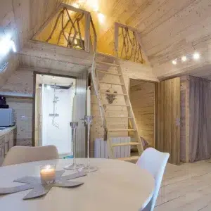 Intérieur du chalet A-Frame Iorik – séjour et mezzanine Intérieur du chalet A-Frame Iorik : coin repas, mezzanine et échelle en bois sous la charpente apparente, à Gérardmer