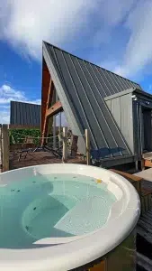Bain nordique privatif du chalet A-Frame Iorik – Gérardmer, Vosges Bain nordique rond en extérieur devant le chalet A-Frame Iorik, terrasse en bois et ciel bleu, près de Gérardmer dans les Vosges
